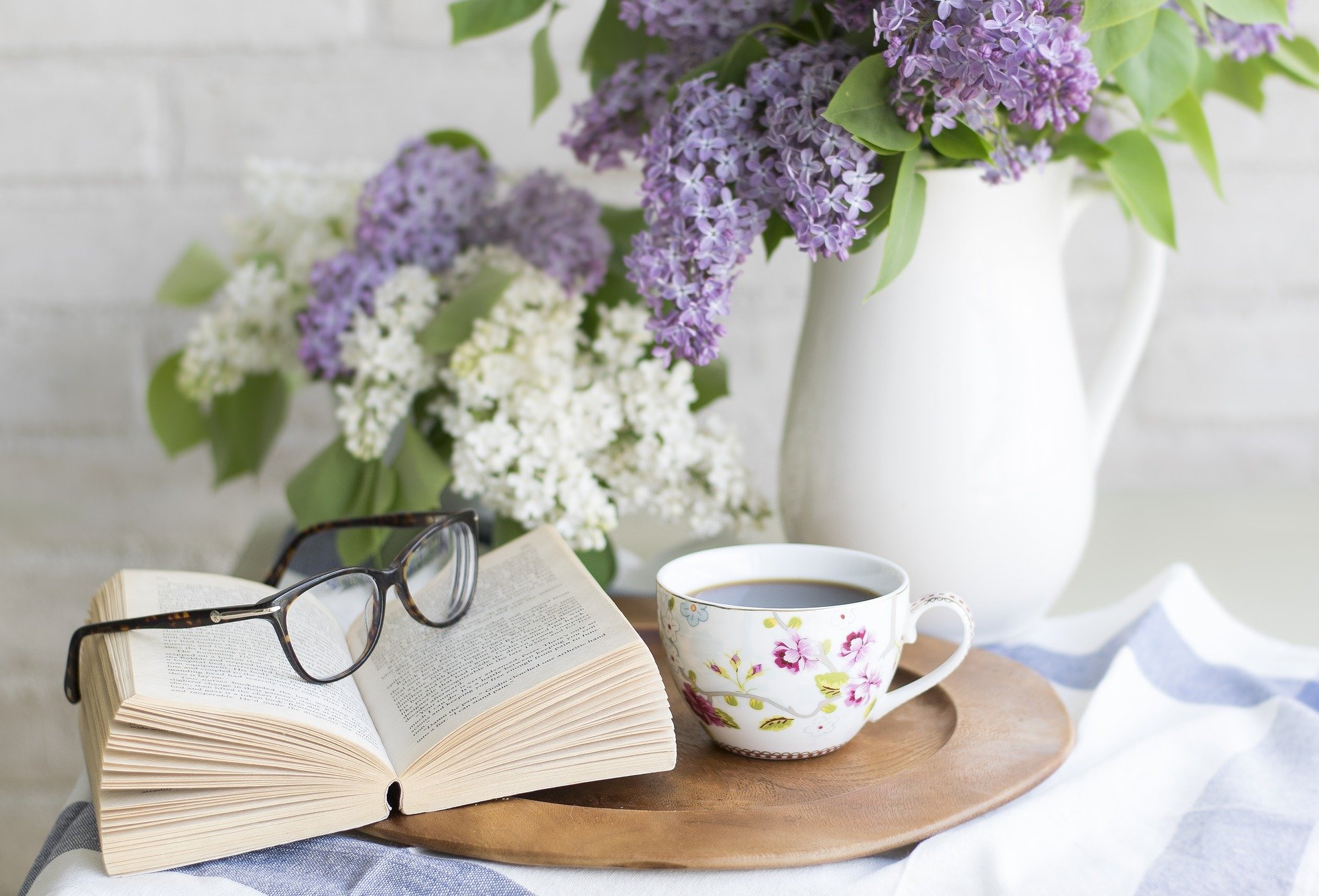 Ein offenen Buch liegt auf dem Tisch, eine Brille obendrauf, eine Tasse und eine Blumenvase stehen rechts daneben.